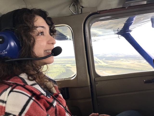 Woman wearing an aviation headset sits inside a small airplane, looking out the window at the landscape below.