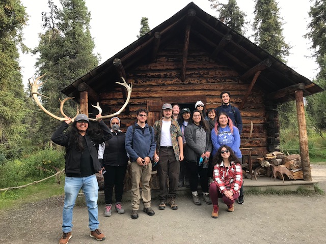 Group of people pose in front of a rustic log cabin in the woods; one person holds a large set of moose antlers over their head.