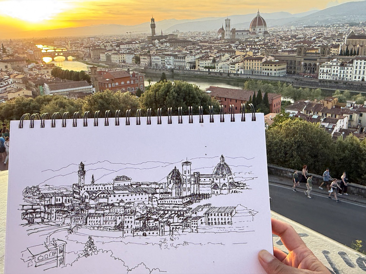 Sketchbook drawing of the Florence skyline held up against the city at sunset, with the Arno River and Florence Cathedral’s dome in the background.