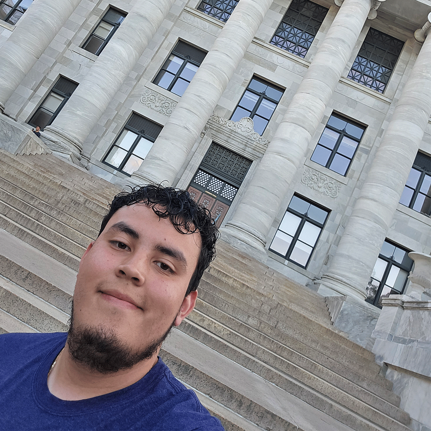 edwin in college A young man smiles for a selfie in front of a large stone building with tall columns and a grand staircase. The architecture suggests a historic or academic institution, possibly a university or government building.