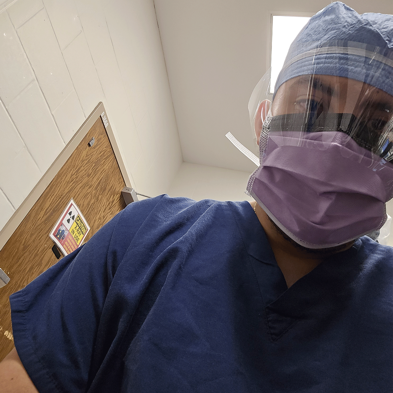 edwin in scrubs A healthcare worker wearing blue scrubs, a purple surgical mask, a face shield, and a blue surgical cap stands in a clinical room. A warning sign for radiation is visible on a wooden door in the background, indicating a medical or diagnostic imaging environment.