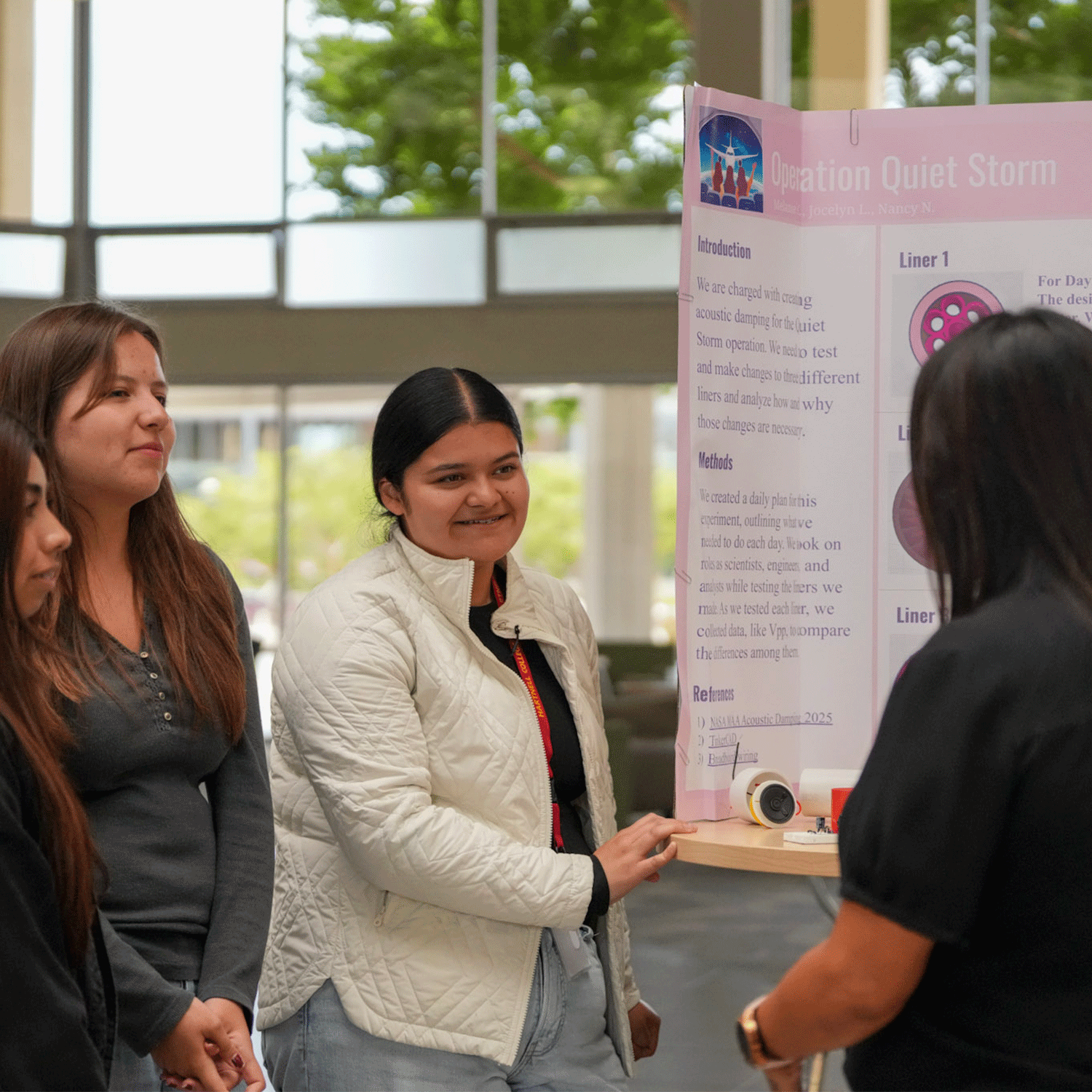NASA MUREP 1 Three high school students present their project titled “Operation Quiet Storm” during the NASA MUREP Aerospace Academy Symposium at Hartnell College. One student gestures toward a tri-fold poster while an adult listens attentively. The event takes place in a bright, modern indoor space with large windows in the background.