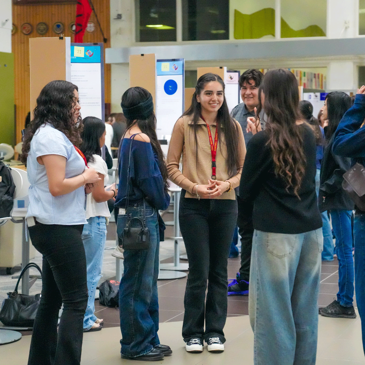 NASA MUREP2 Groups of students engage in conversation during the NASA MUREP Aerospace Academy Symposium at Hartnell College. One student wearing a tan sweater and red lanyard stands at the center, smiling while speaking to peers. Poster boards with student projects are displayed in the background inside a bright, open space.