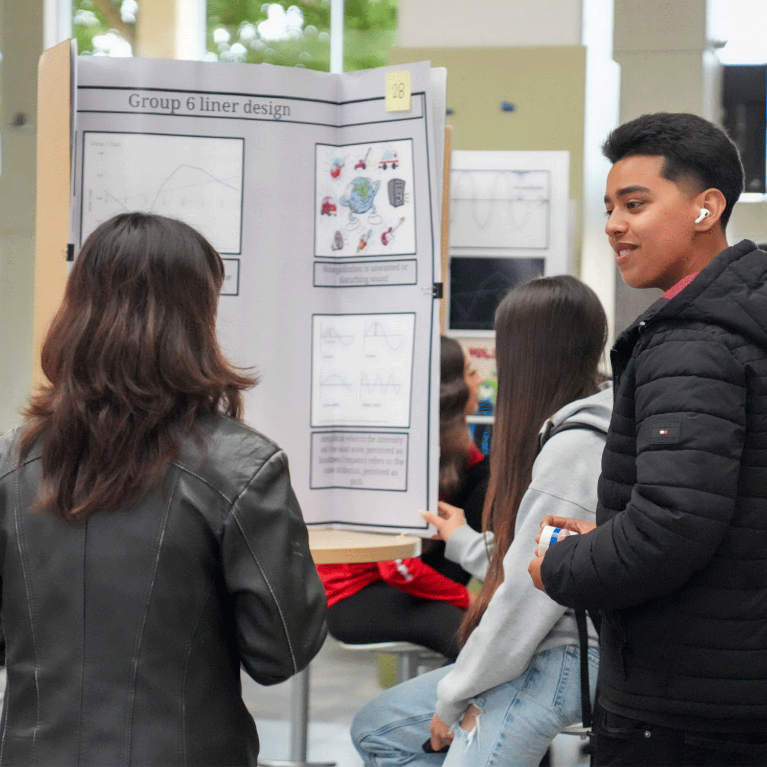maa3 Three students stand around a trifold presentation board labeled "Group 6 liner design" at a science or engineering fair. The display includes graphs, diagrams, and colorful illustrations. One student, wearing a black puffer jacket and AirPods, smiles while speaking. Another student in a grey hoodie leans against the table, and a third, with long brown hair and a black leather jacket, faces the board. The event is taking place in a bright, modern indoor space with large windows and other seated participants in the background.