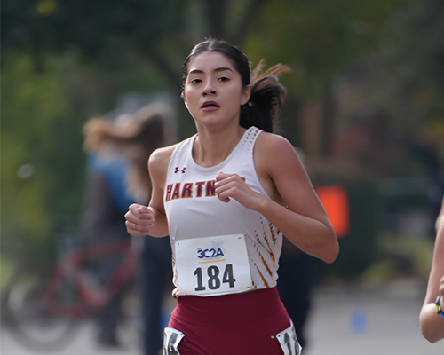 Hartnell College women’s cross country runner in a white and maroon uniform running on a paved course during a race.