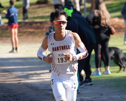 A focused male cross country runner, number 193, from Hartnell College, races outdoors. He is wearing a white singlet with brown trim, sunglasses, and arm sleeves.