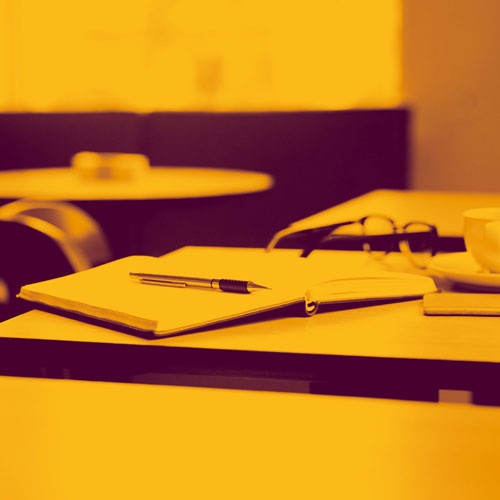 Open notebook and pen on a classroom table, with chairs and other learning materials in the background, shown in a warm yellow and purple color palette.