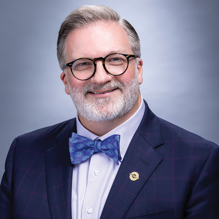Headshot of Dr. Russell Lowery-Hart smiling, wearing glasses, a navy suit, and a blue patterned bow tie.