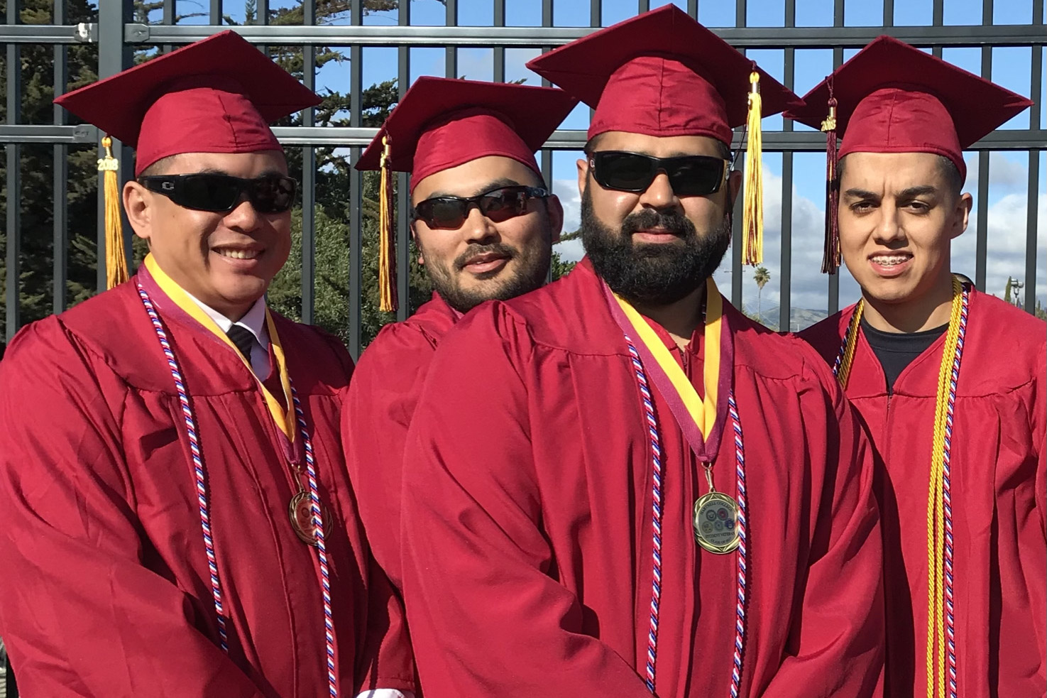 Four graduates in red caps and gowns with sunglasses, wearing medals and cords, smiling outdoors.