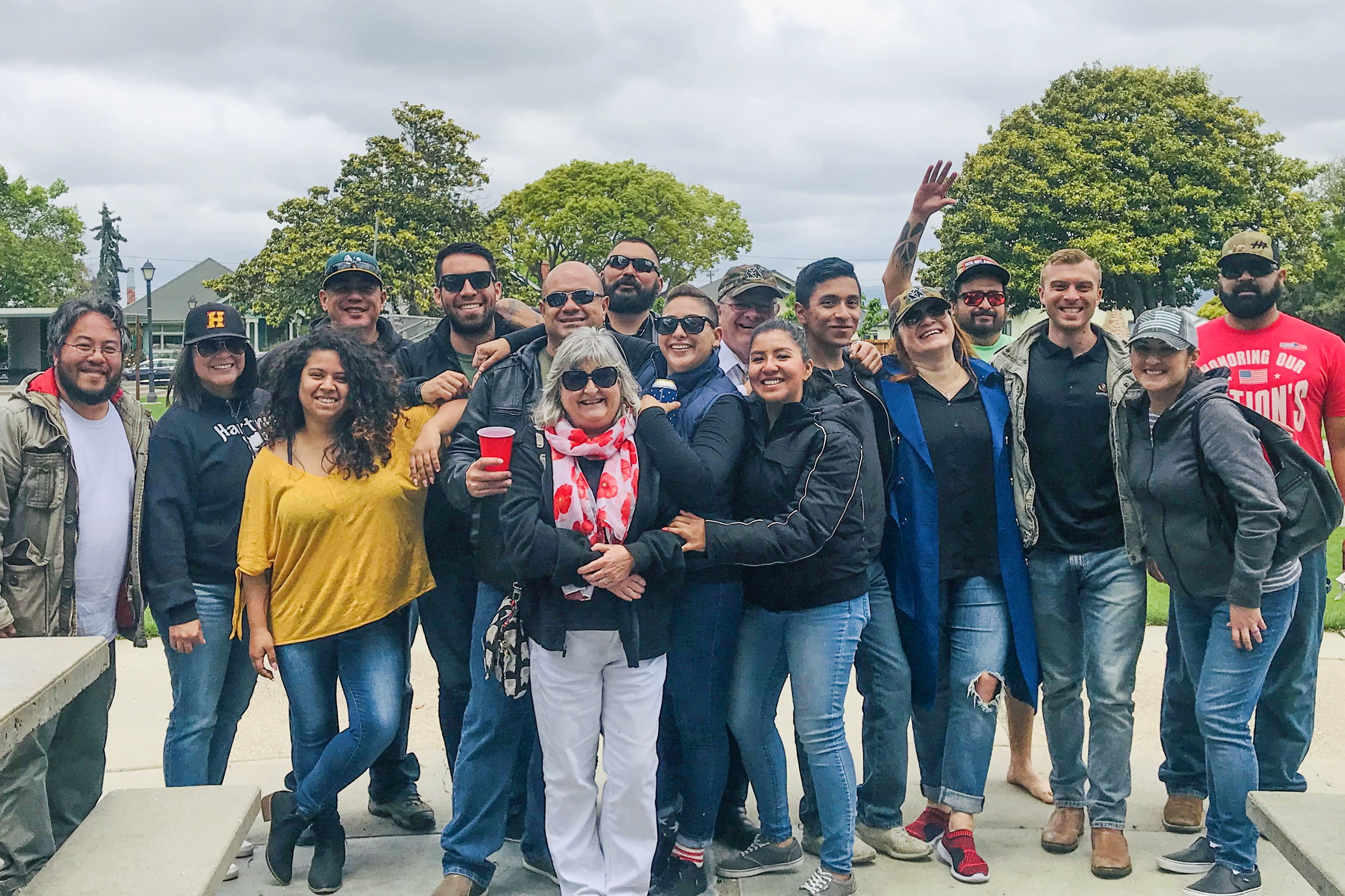 Group of sixteen people outdoors, smiling and posing together, some wearing sunglasses and hats, with trees and cloudy sky in the background.