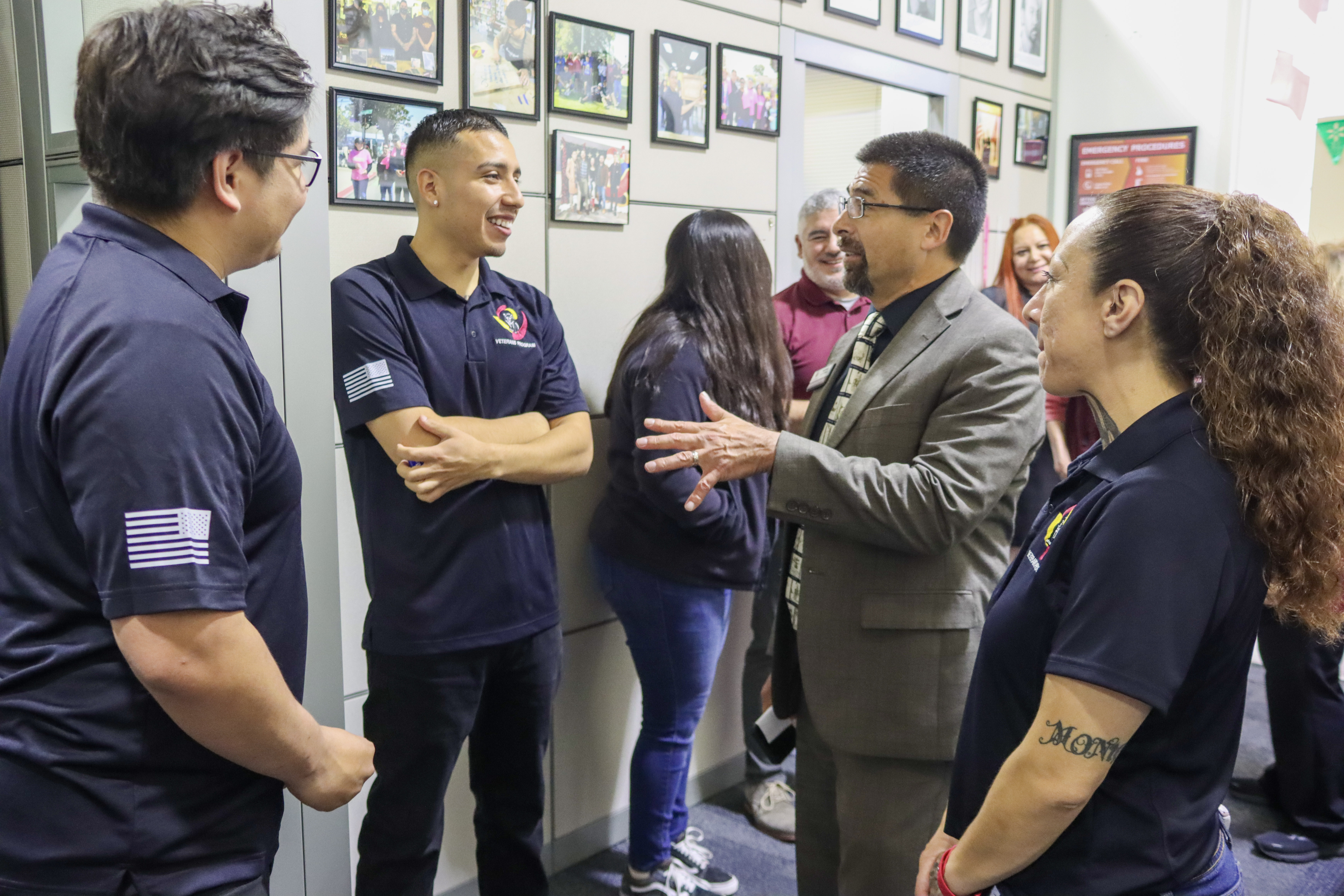 Group of people indoors, with three in matching navy polo shirts talking to a man in a suit, while others listen in the background.