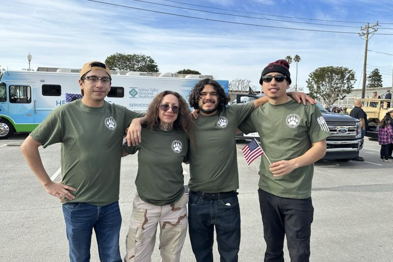 Four people wearing matching green shirts with a paw print logo stand arm-in-arm outdoors, one holding a small U.S. flag.