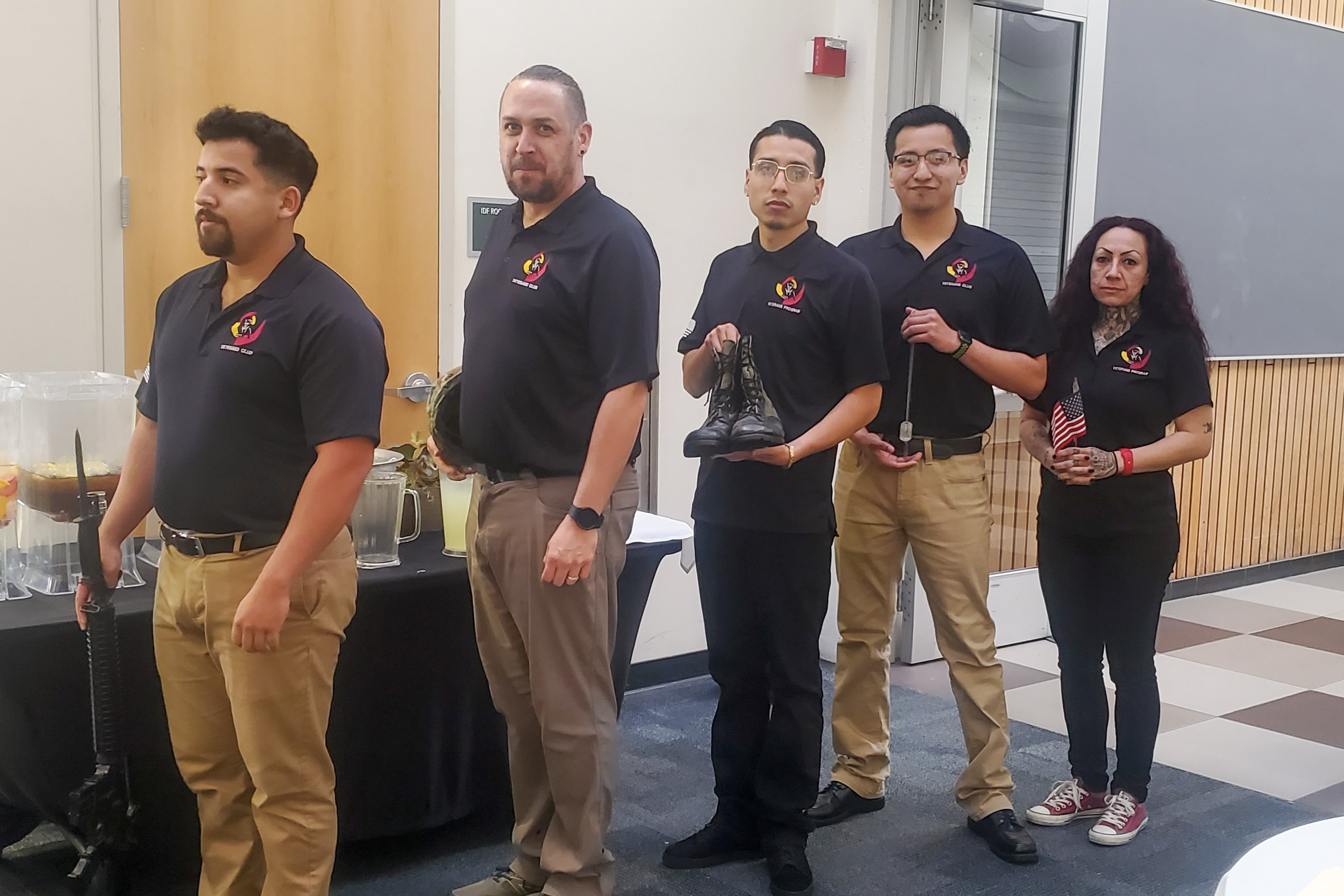 Five people in matching black polo shirts and khaki or black pants standing in a line indoors, each holding symbolic military items including a rifle, helmet, boots, dog tags, and a small American flag.