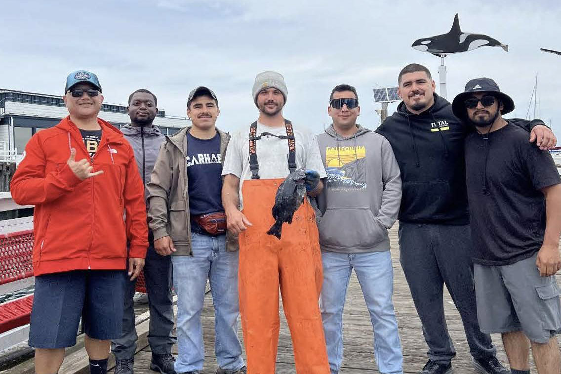 Seven men on a dock, one holding a freshly caught fish, with a marina and orca sculpture in the background.