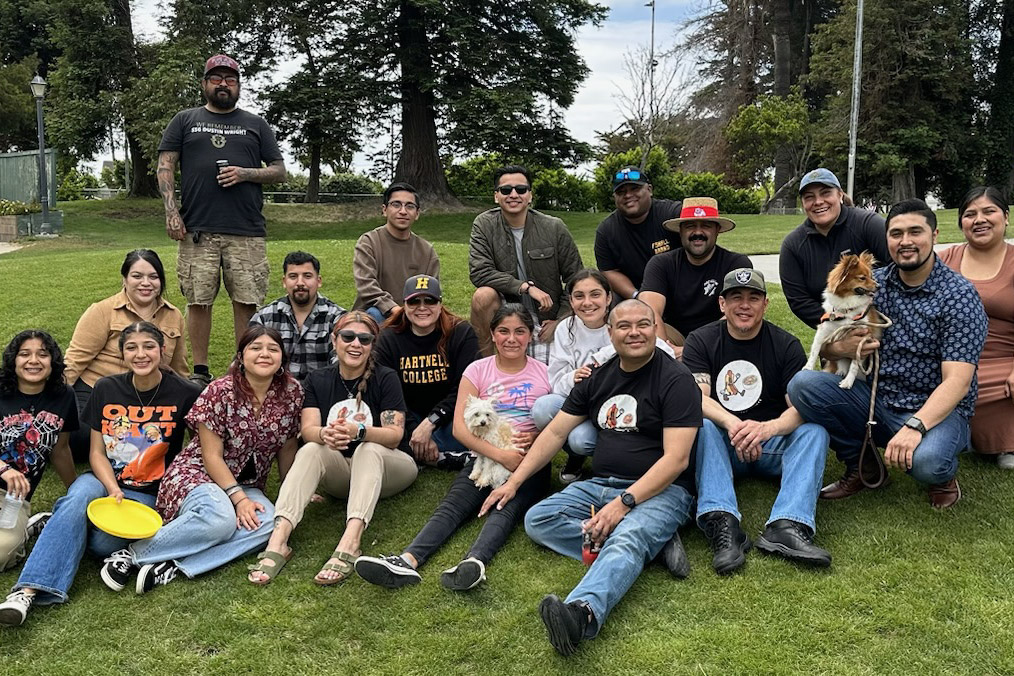 Large group sitting on a grassy park lawn with a dog in the front row during an outdoor social gathering.